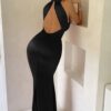 Brunette lady with hair in bun in open back black satin maxi dress leaning on shelf with hand under chin.